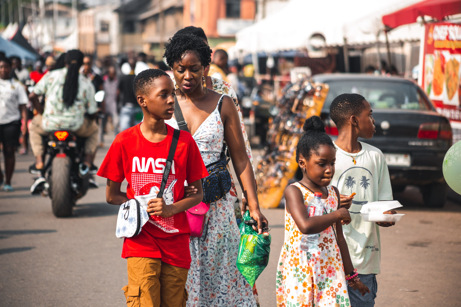 A woman carrying bags walks beside three children along a busy street market.