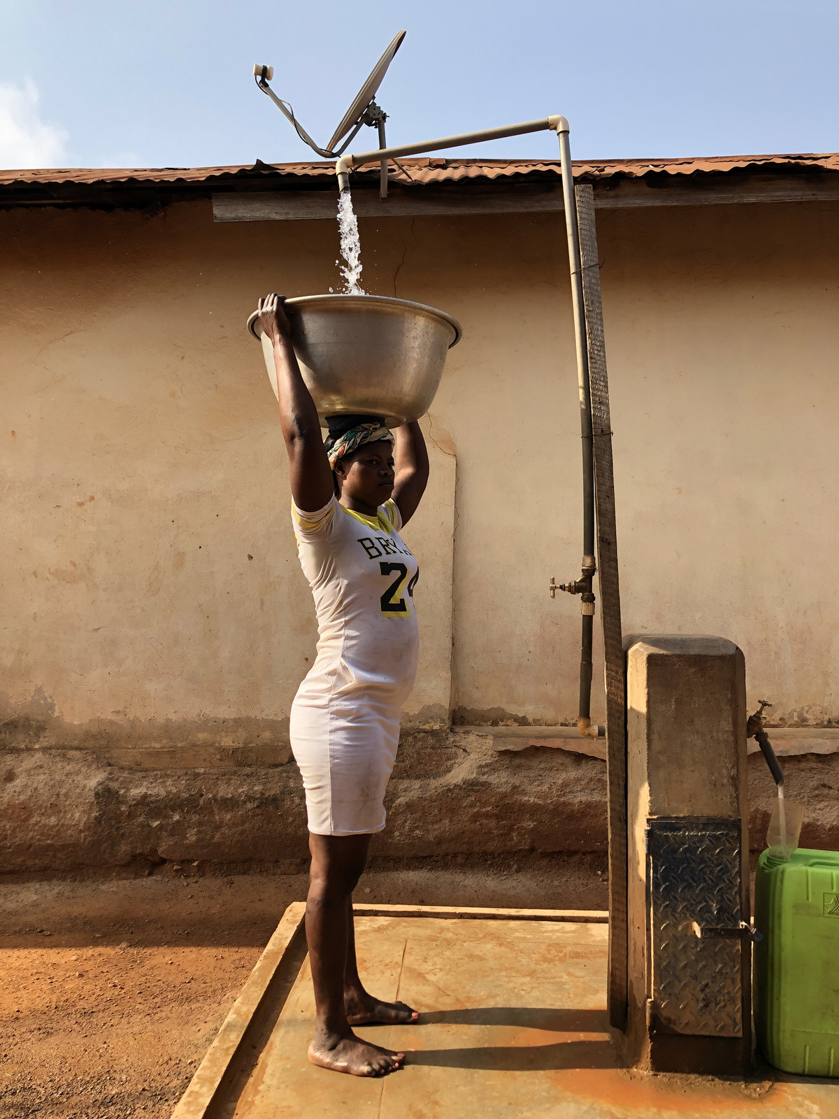 A woman steadies a metal basin on her head as water flows into it from an overhead standpipe.