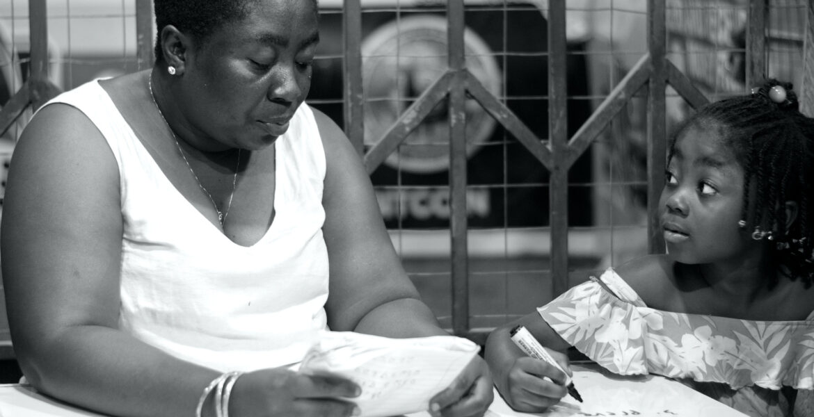 Black-and-white photograph of a woman holding an open exercise book beside a child holding a pen over a workbook.