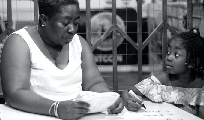 Black-and-white photograph of a woman holding an open exercise book beside a child holding a pen over a workbook.