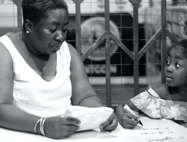 Black-and-white photograph of a woman holding an open exercise book beside a child holding a pen over a workbook.