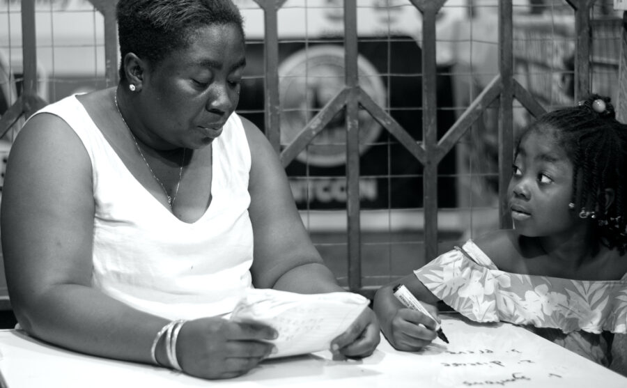 Black-and-white photograph of a woman holding an open exercise book beside a child holding a pen over a workbook.