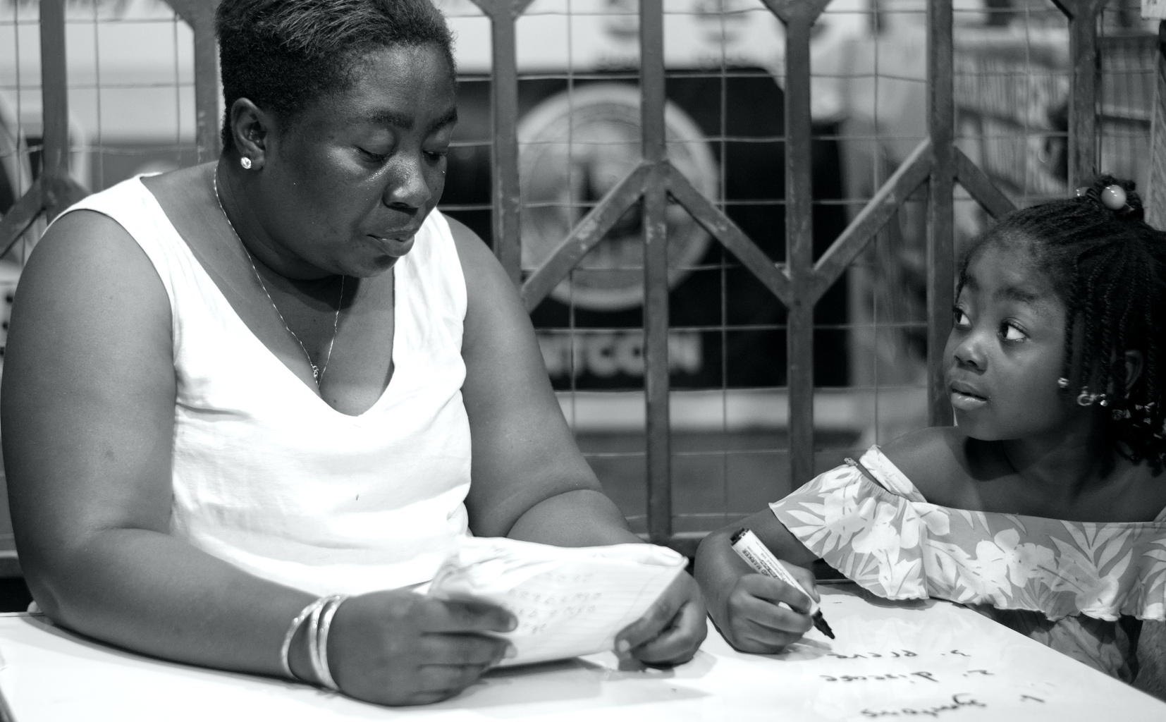 Black-and-white photograph of a woman holding an open exercise book beside a child holding a pen over a workbook.