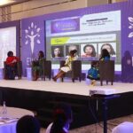 Four women seated on stage during a panel discussion against a purple EWN backdrop.